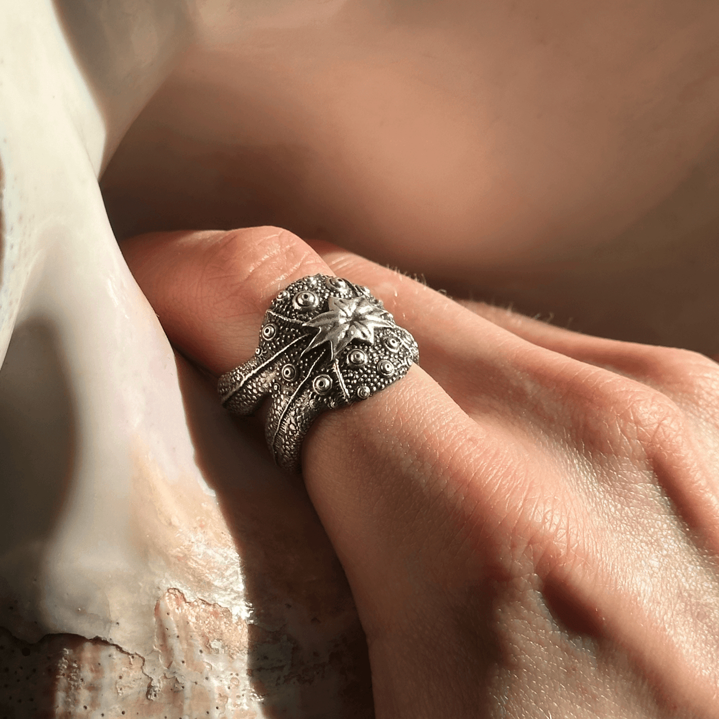 A model's hand adorned with the Beast ring, featuring a bold, starfish-inspired design with raised textures and organic detailing. The sunlight highlights the intricate craftsmanship against a neutral seashell background.