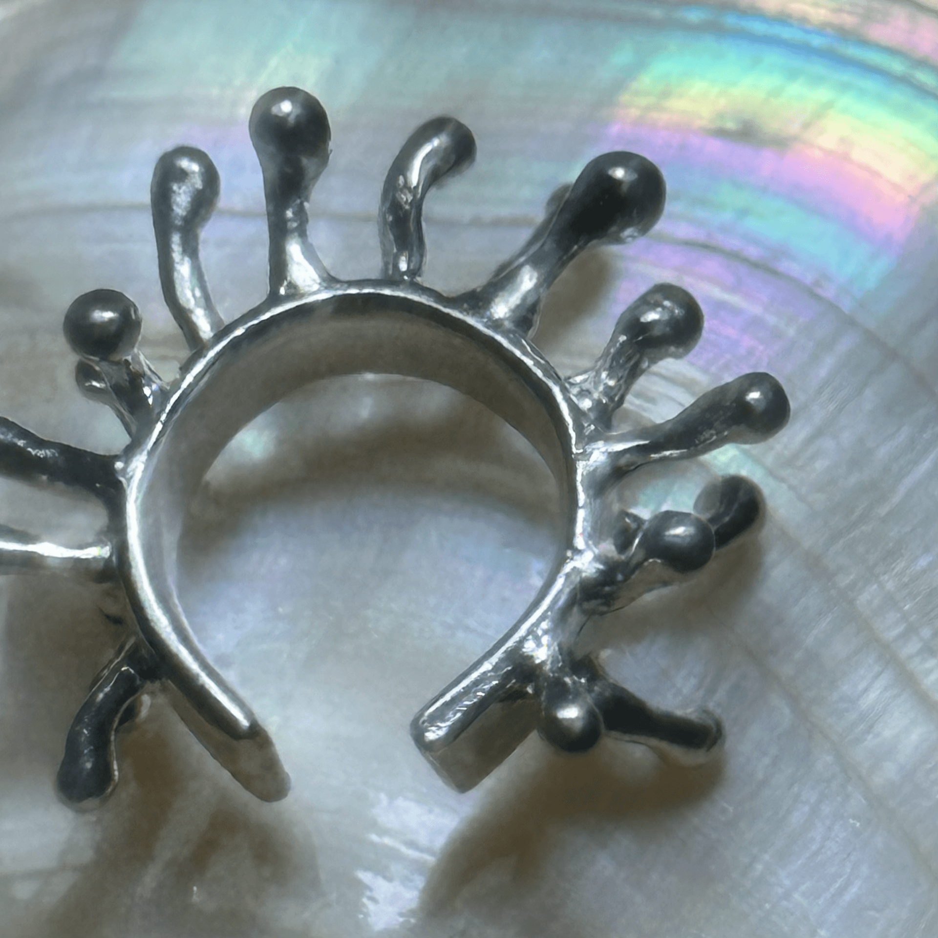 A detailed macro shot of a silver Hydra earcuff resting on an iridescent shell surface. The polished metal reflects light, highlighting its intricate organic texture.