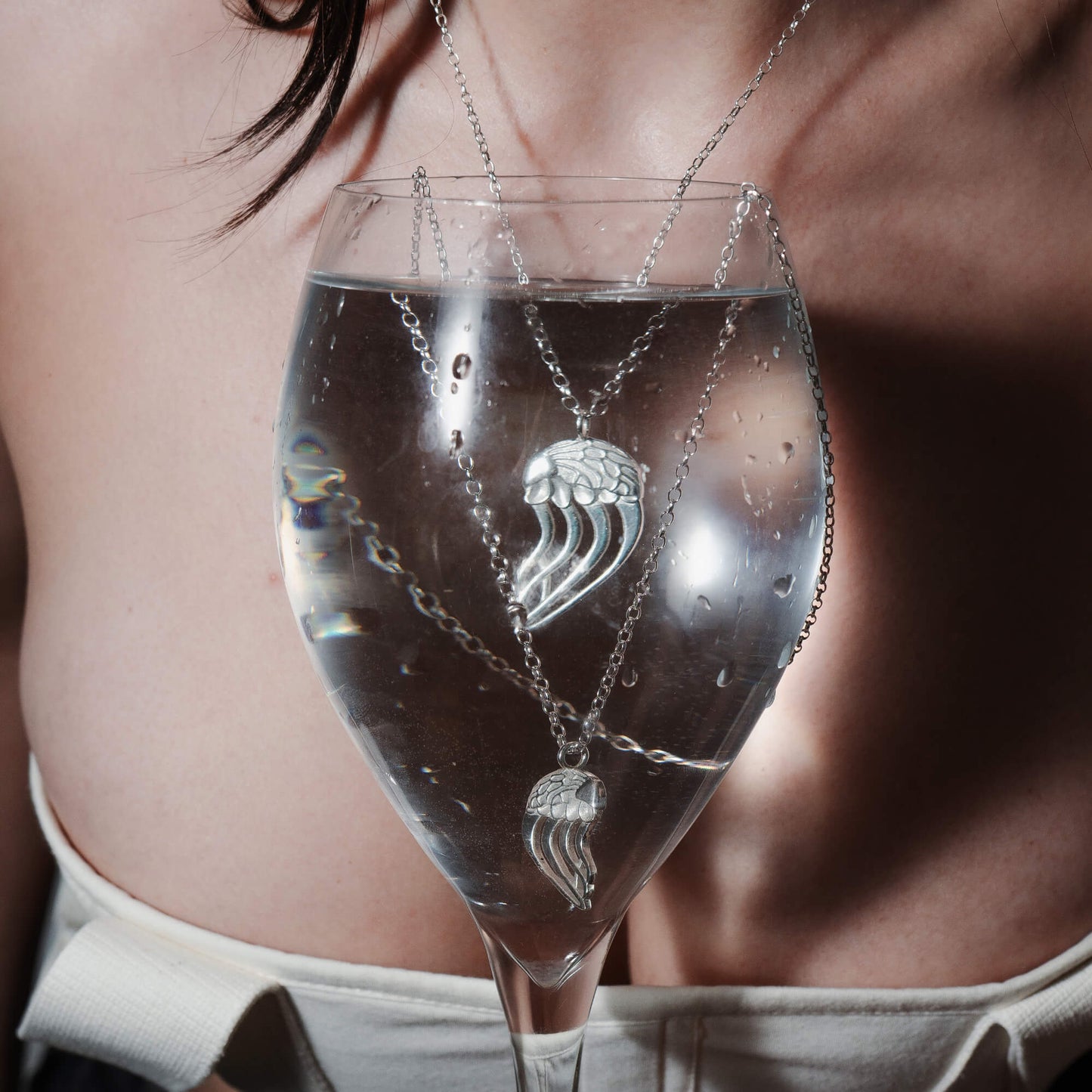 The Jellyfish necklace, with its intricate silver design, is submerged in a glass of water, creating an ethereal and reflective effect. The pendant's delicate tentacle-like elements are beautifully visible through the water.