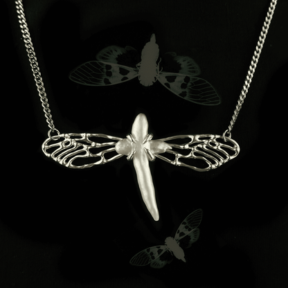 Close-up of the Metamorphosis necklace featuring a silver insect-like design with intricately crafted wings and a central pearl. The pendant symbolizes transformation, set against a dark background with faint insect imagery.