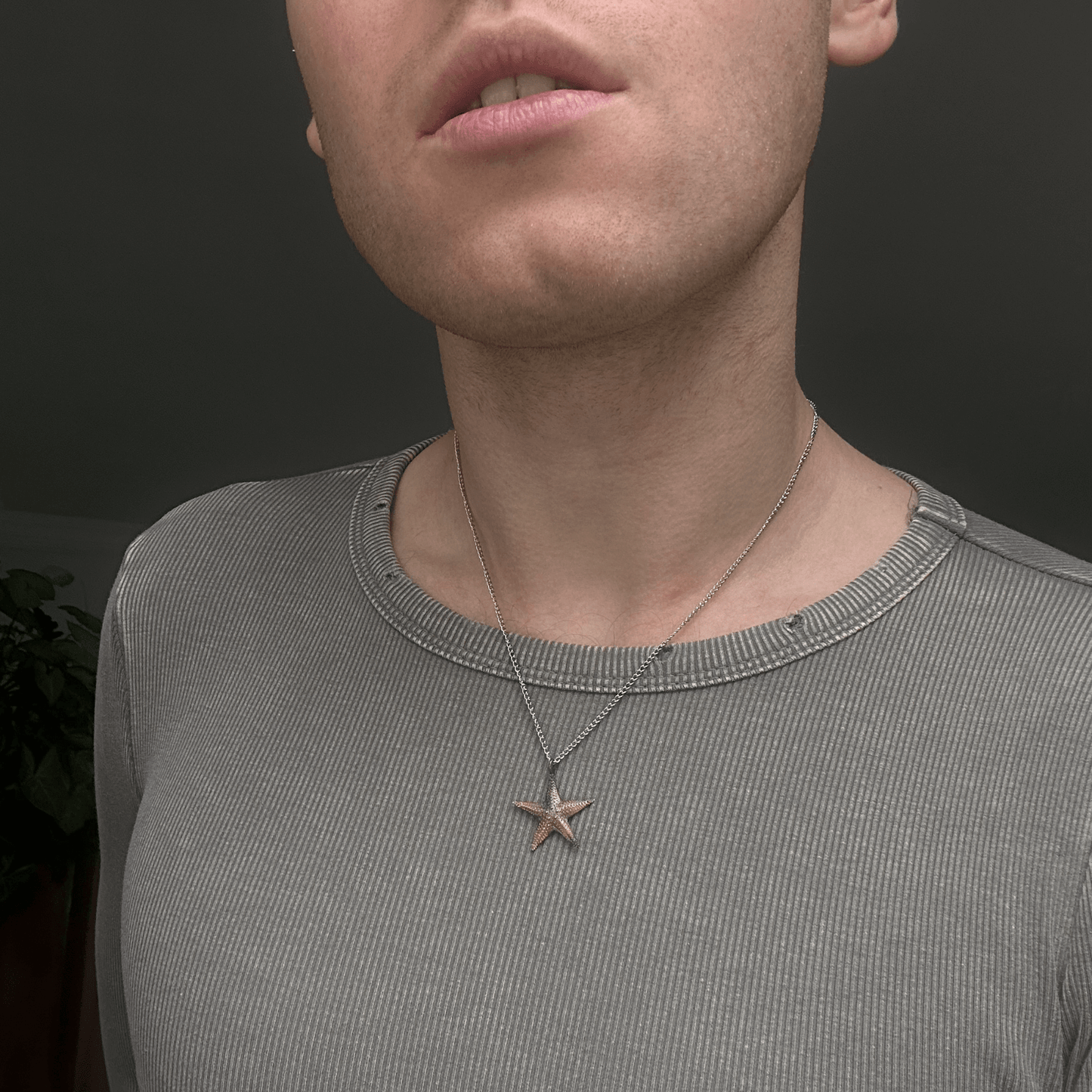 A silver starfish pendant necklace worn on a thin chain, showcased on a model wearing a ribbed gray shirt. The pendant's textured design is inspired by marine starfish species, adding an organic touch to the minimal aesthetic.