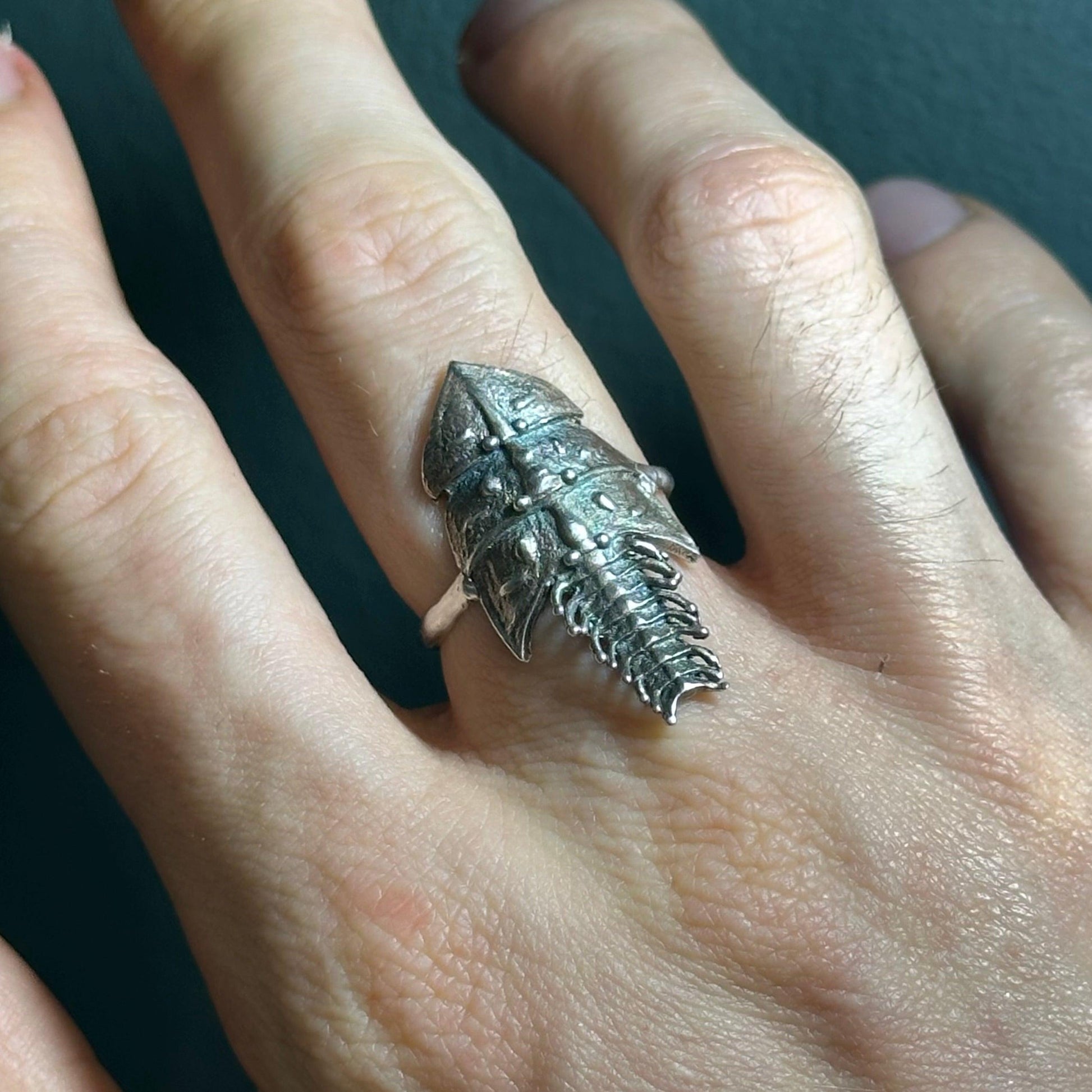 A close-up of a hand wearing the Trilobite ring, highlighting its detailed, segmented armor-like design. The textured silver surface reflects light, emphasizing its insect-inspired aesthetic.