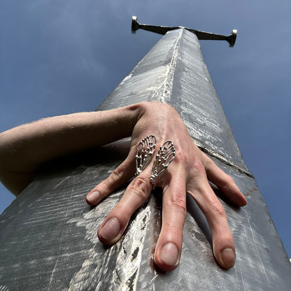 The Wings Ring showcased outdoors, worn on a hand grasping a metallic structure under a bright blue sky. The natural light emphasizes the fine details of the cicada wing-inspired design.